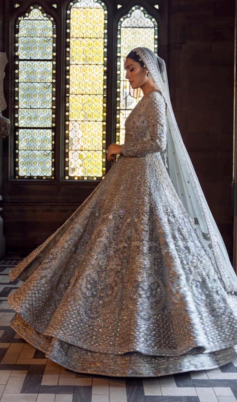 May include: A silver, long-sleeved wedding gown with intricate beading and embroidery. The dress features a full skirt and a long, flowing veil. The gown is worn by a person standing in front of a large window with stained glass.