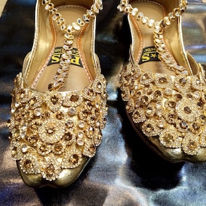 May include: A pair of gold-colored, embellished sandals. The sandals feature intricate floral patterns and are adorned with sparkling rhinestones. The straps and upper are covered in decorative elements, creating a glamorous look. The soles are also gold.