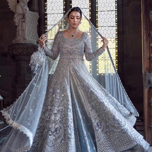 May include: A silver-gray bridal gown with intricate embroidery and embellishments. The long-sleeved dress features a square neckline and a flowing skirt. A sheer veil and matching beaded purse complete the elegant ensemble. The background includes a stained-glass window.