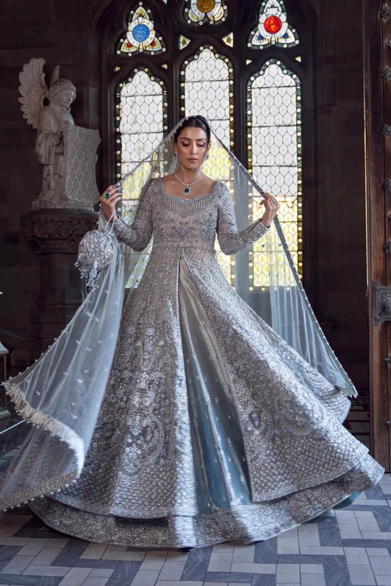 May include: A silver-gray bridal gown with intricate embroidery and embellishments. The long-sleeved dress features a square neckline and a flowing skirt. A sheer veil and matching beaded purse complete the elegant ensemble. The background includes a stained-glass window.