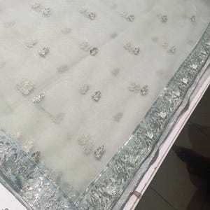 May include: A sheer, light blue fabric with delicate silver floral embroidery. The edge is trimmed with a silver and light blue decorative border. The fabric is folded on a white surface. The overall theme is bridal or formal wear.