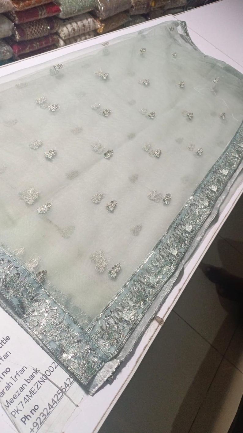 May include: A sheer, light blue fabric with delicate silver floral embroidery. The edge is trimmed with a silver and light blue decorative border. The fabric is folded on a white surface. The overall theme is bridal or formal wear.