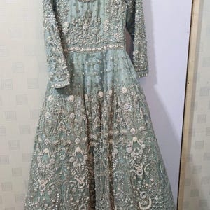 May include: A long, sage green formal gown with intricate floral embroidery and beadwork. The dress features long sleeves, a round neckline, and a full skirt. The gown is hanging on a metal hanger against a white background.