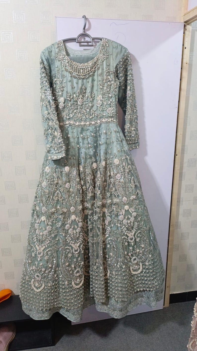 May include: A long, sage green formal gown with intricate floral embroidery and beadwork. The dress features long sleeves, a round neckline, and a full skirt. The gown is hanging on a metal hanger against a white background.