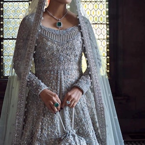 May include: A silver-gray embellished gown with long sleeves, a matching veil, and a beaded purse. The dress features intricate beadwork and a square neckline. The ensemble includes a necklace and ring with emerald-colored stones. The background has a stained-glass window.