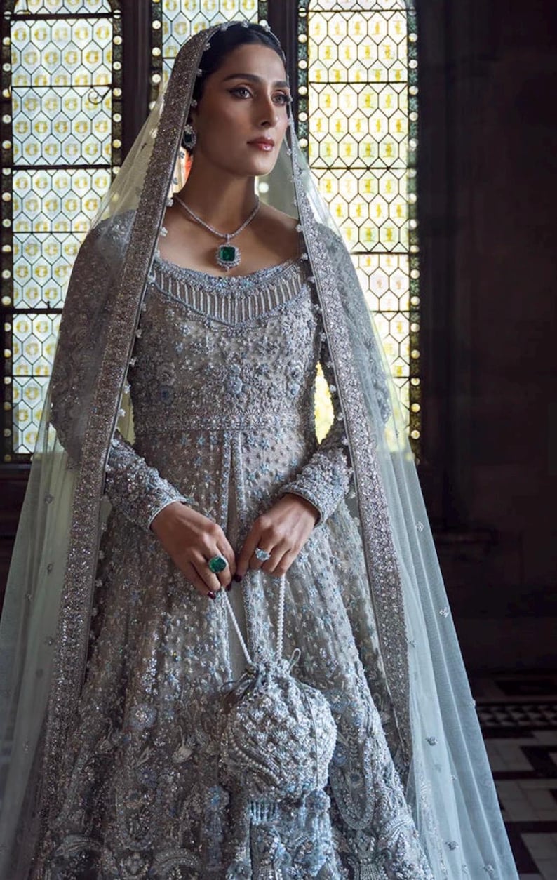 May include: A silver-gray embellished gown with long sleeves, a matching veil, and a beaded purse. The dress features intricate beadwork and a square neckline. The ensemble includes a necklace and ring with emerald-colored stones. The background has a stained-glass window.