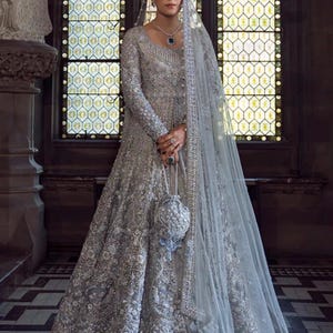 May include: A silver-gray embellished gown with long sleeves and a matching veil. The dress features intricate beadwork and a small, beaded purse. The background includes a stained-glass window and stone architecture.