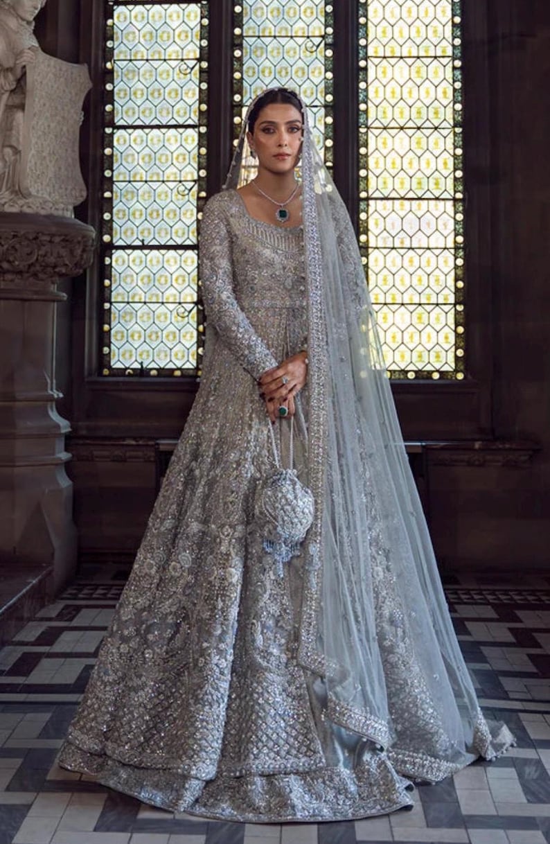 May include: A silver-gray embellished gown with long sleeves and a matching veil. The dress features intricate beadwork and a small, beaded purse. The background includes a stained-glass window and stone architecture.