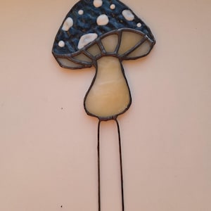 May include: A stained glass mushroom garden stake. The mushroom cap is dark blue with white spots, and the stem is a light cream color. Two metal rods extend from the bottom for insertion into the ground. The mushroom is approximately 6 inches tall.