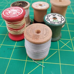 May include: A collection of five vintage wooden thread spools. One red spool holds beige thread with an Aunt Lydia's label. A central spool features grey thread and a Belding Corticelli label. Other spools include natural wood and green finishes.