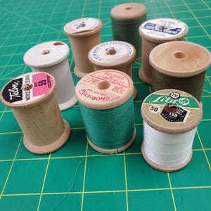 May include: A collection of nine vintage wooden sewing thread spools in various colors including white, green, and brown. Each spool features a circular paper label on top with printed brand names like Talon, Belding Corticelli, and Lily.