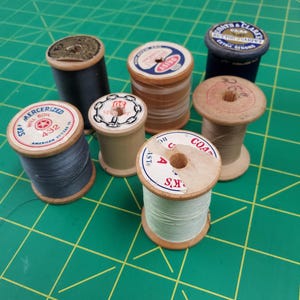 May include: A collection of vintage wooden spools of thread in various colors, including black, blue, and beige. Some spools have labels with text like "MERCERIZED" and "COATS & CLARKS". The spools are arranged on a green grid surface.