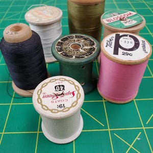 May include: A collection of vintage thread spools in various colors, including black, white, pink, and green. The spools are made of wood and cardboard, with labels indicating thread type and brand. The spools are arranged on a green cutting mat.