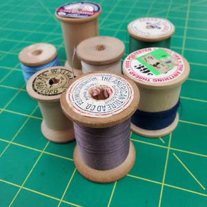 May include: A collection of seven vintage wooden spools of thread in various colors including purple, dark blue, light blue, white, and green. Each spool features a circular paper label on top with printed brand text such as The American Thread Co.