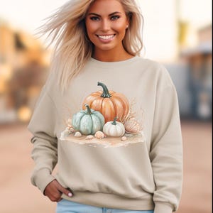 May include: Beige sweatshirt with a watercolour design of pumpkins, seashells, and sand. The design includes an orange, a light blue, and a white pumpkin. The text "Gildan 18000 Gand" is printed at the bottom of the sweatshirt.