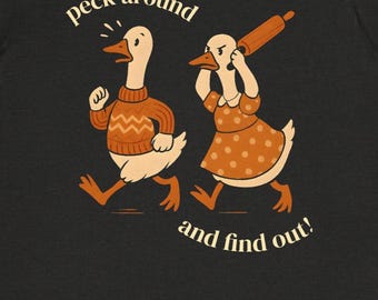 May include: Dark gray t-shirt featuring a cartoon illustration of two geese. One goose is wearing an orange sweater and running, while the other goose is holding a rolling pin. The text "peck around and find out!" is arched above and below the geese.