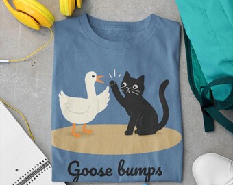 May include: A blue t-shirt featuring a cartoon duck and a black cat giving each other a high-five. The text "Goose bumps" is printed below the illustration. Yellow headphones, a green backpack, white sneakers, and colorful markers are also visible.