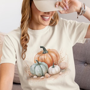 May include: A cream-coloured t-shirt with a watercolour design of pumpkins and seashells. The pumpkins are orange, blue-green, and white, with a black stem. Seashells and dried grass surround the pumpkins.