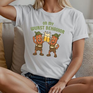May include: White Bella Canvas 3001 t-shirt with a cartoon of two sausages in lederhosen toasting with beer. The text "on my WURST BEHAVIOR" is printed above the cartoon. The shirt is white.