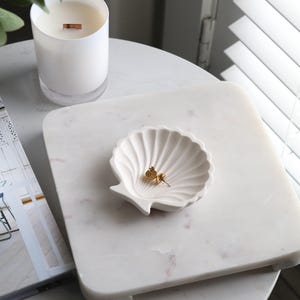 May include: A white marble tray with a seashell-shaped dish holding gold earrings. A white candle with a wooden wick sits nearby. The items are on a white table, with a magazine and window blinds in the background.