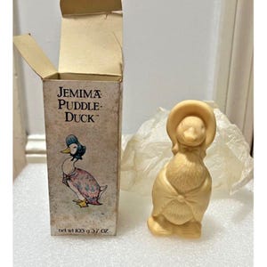 Puede incluir: Una figura de Jemima Puddle-Duck con su caja. La figura es de color beige claro y lleva un gorro y un chal. La caja tiene las palabras "Jemima Puddle-Duck" y una ilustración del pato. La caja también indica el peso neto de 105 g.