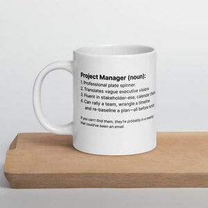 Puede incluir: Taza de cerámica blanca con el texto "Project Manager (noun):" seguido de una definición humorística. La taza está sobre una superficie de madera. El texto incluye frases como "Professional plate spinner" y "all before lunch."