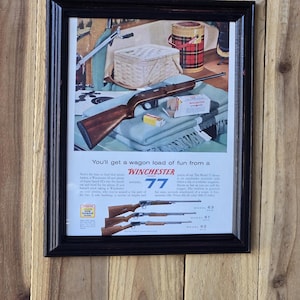May include: Framed vintage advertisement for Winchester firearms. The ad features a rifle, a picnic basket, and a thermos. Text reads "You'll get a wagon load of fun from a Winchester". The frame is black, and the background is wood.
