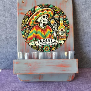 May include: A gray wooden tequila shot glass holder with a colorful circular graphic featuring a skeleton in a sombrero holding a shot glass. The graphic includes the text "TEQUILA SERVED HERE" and a tequila bottle. The holder has a shelf with five shot glasses.