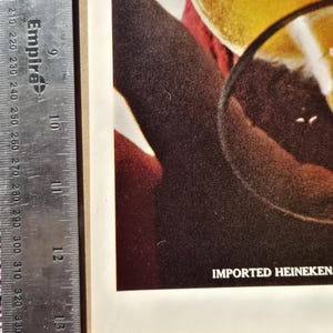 Pu&ograve; includere: Primo piano di una pubblicit&agrave; vintage con un bicchiere di birra e una fetta di limone. Il testo "IMPORTED HEINEKEN" &egrave; visibile. L'immagine ha una calda tavolozza di colori, incentrata sulla birra e sul bicchiere.