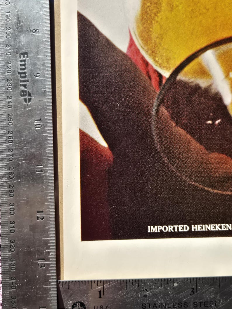 Pu&ograve; includere: Primo piano di una pubblicit&agrave; vintage con un bicchiere di birra e una fetta di limone. Il testo "IMPORTED HEINEKEN" &egrave; visibile. L'immagine ha una calda tavolozza di colori, incentrata sulla birra e sul bicchiere.