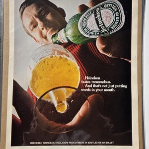 Pu&ograve; includere: Annuncio vintage con una persona che beve birra Heineken da una bottiglia verde in un bicchiere. La bottiglia ha un'etichetta bianca e rossa. Il testo recita: "Heineken ha un gusto tremendo. E non si tratta solo di mettere parole in bocca."
