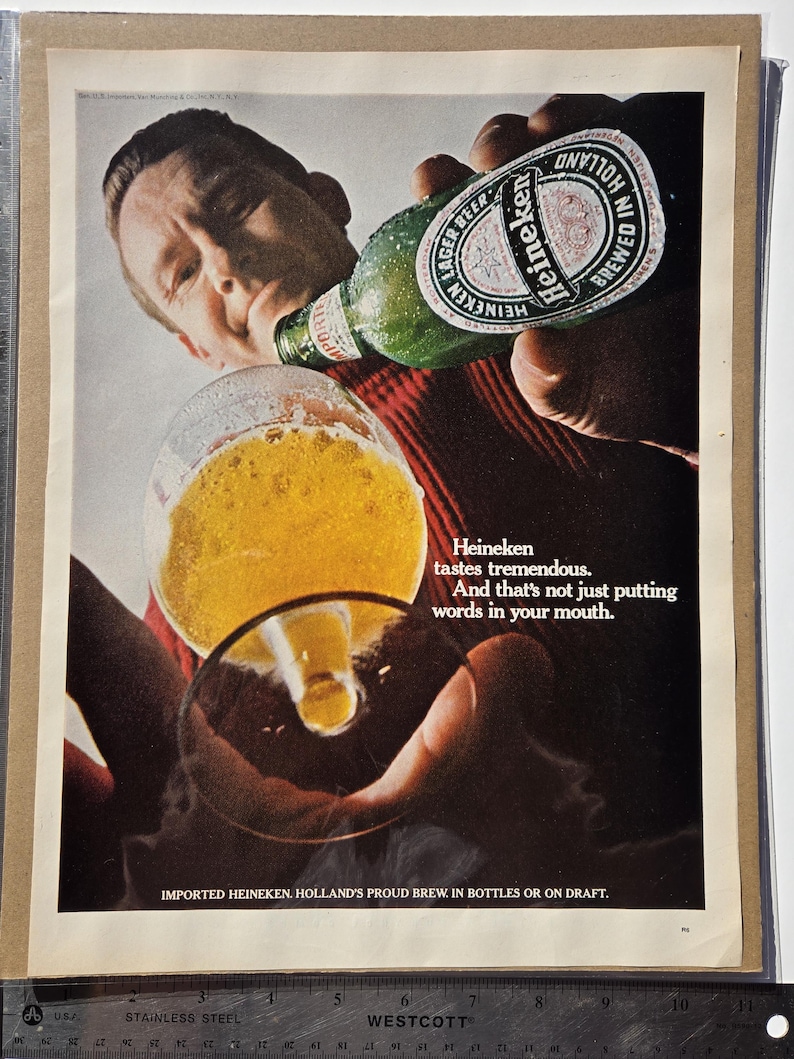Pu&ograve; includere: Annuncio vintage con una persona che beve birra Heineken da una bottiglia verde in un bicchiere. La bottiglia ha un'etichetta bianca e rossa. Il testo recita: "Heineken ha un gusto tremendo. E non si tratta solo di mettere parole in bocca."