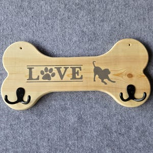 May include: Wooden dog bone-shaped wall hook with the word "LOVE" and a paw print and dog silhouette. The light-colored wood has two black hooks and is designed for pet accessories.