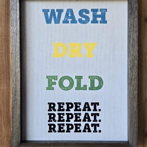 May include: Wooden framed sign with the words "WASH DRY FOLD" in blue, yellow, and green, followed by "REPEAT. REPEAT. REPEAT." in black. The sign is set against a white background.