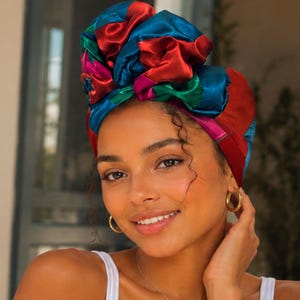 Può includere: Una donna indossa un cappuccio di raso colorato davanti a uno specchio. Il cappuccio presenta un vivace mix di tonalità rosse, blu, verdi e rosa. Indossa una canotta bianca con la scritta "THE SATIN BONNET".