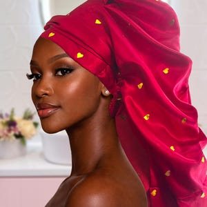 Puede incluir: Un gorro de satén rojo vibrante adornado con pequeños detalles dorados en forma de corazón. El gorro está diseñado para proteger el cabello y mantener los peinados. La tela tiene un brillo lustroso, y el diseño es funcional y elegante.