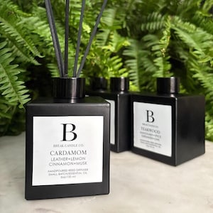 Peut inclure: Diffuseurs à roseaux carrés noirs avec des roseaux noirs. Le diffuseur avant porte une étiquette blanche avec la lettre "B" et les mots "CARDAMOM LEATHER+LEMON CINNAMON+MUSK". L'étiquette indique également "HANDPOURED REED DIFFUSER SMALL BATCH/ESSENTIAL OIL 150 ml".