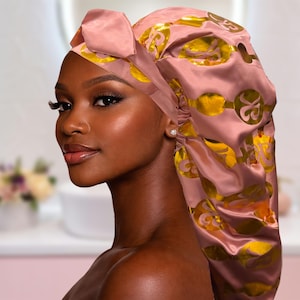 Puede incluir: Un gorro de satén en color rosa empolvado con detalles decorativos dorados. El gorro tiene un lazo en la parte superior y está diseñado para proteger el cabello. El fondo es un interior suave y borroso.