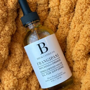 May include: A clear glass bottle with a black dropper top, labeled "FRANGIPANI OIL FOR DIFFUSING." The label includes the brand name "Break Candle Co." and lists ingredients. The bottle contains 4 oz / 120 ml of oil and rests on a textured, orange fabric.