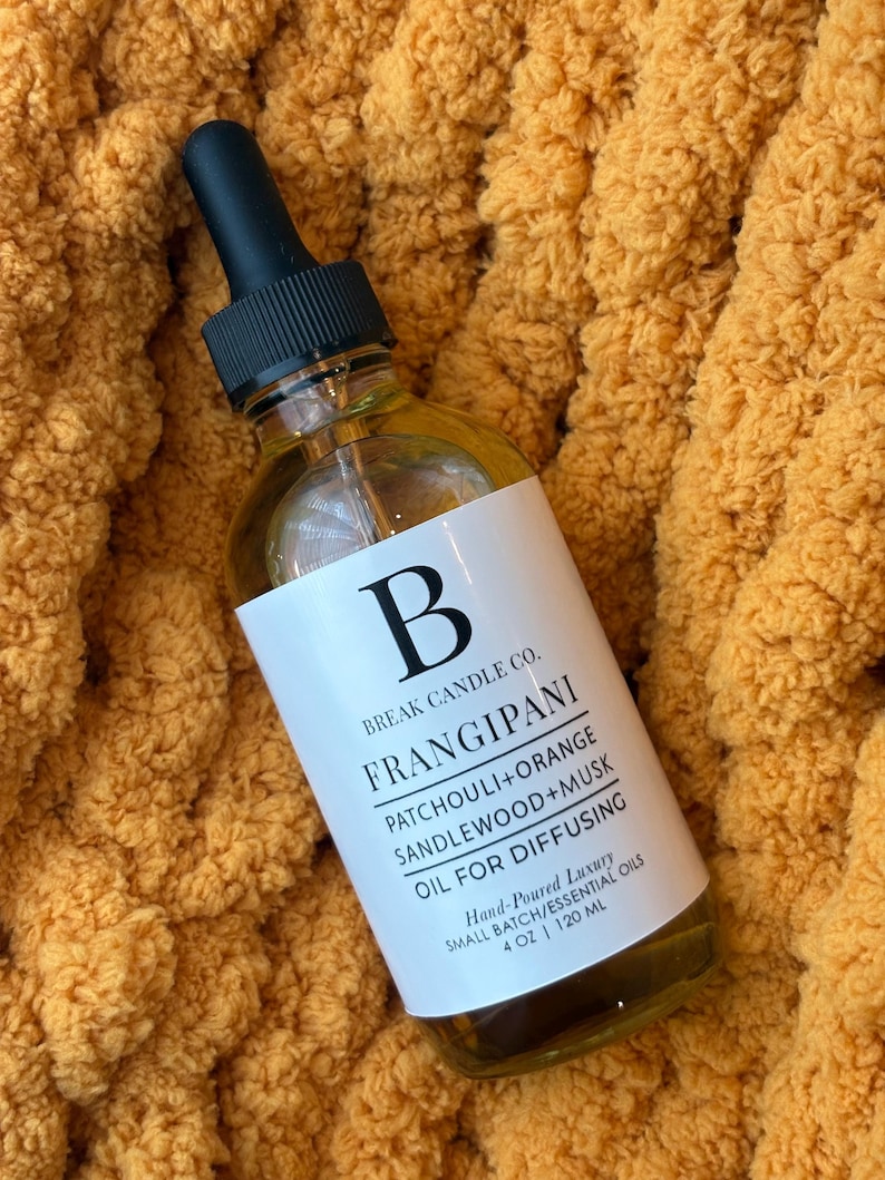 May include: A clear glass bottle with a black dropper top, labeled "FRANGIPANI OIL FOR DIFFUSING." The label includes the brand name "Break Candle Co." and lists ingredients. The bottle contains 4 oz / 120 ml of oil and rests on a textured, orange fabric.