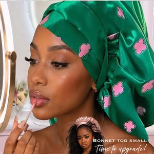 Puede incluir: Un gorro de satén verde con diseños rosas en forma de flor. Una persona se aplica brillo labial mientras usa el gorro. El texto "BONNET TOO SMALL? Time to upgrade!" es visible en la imagen.