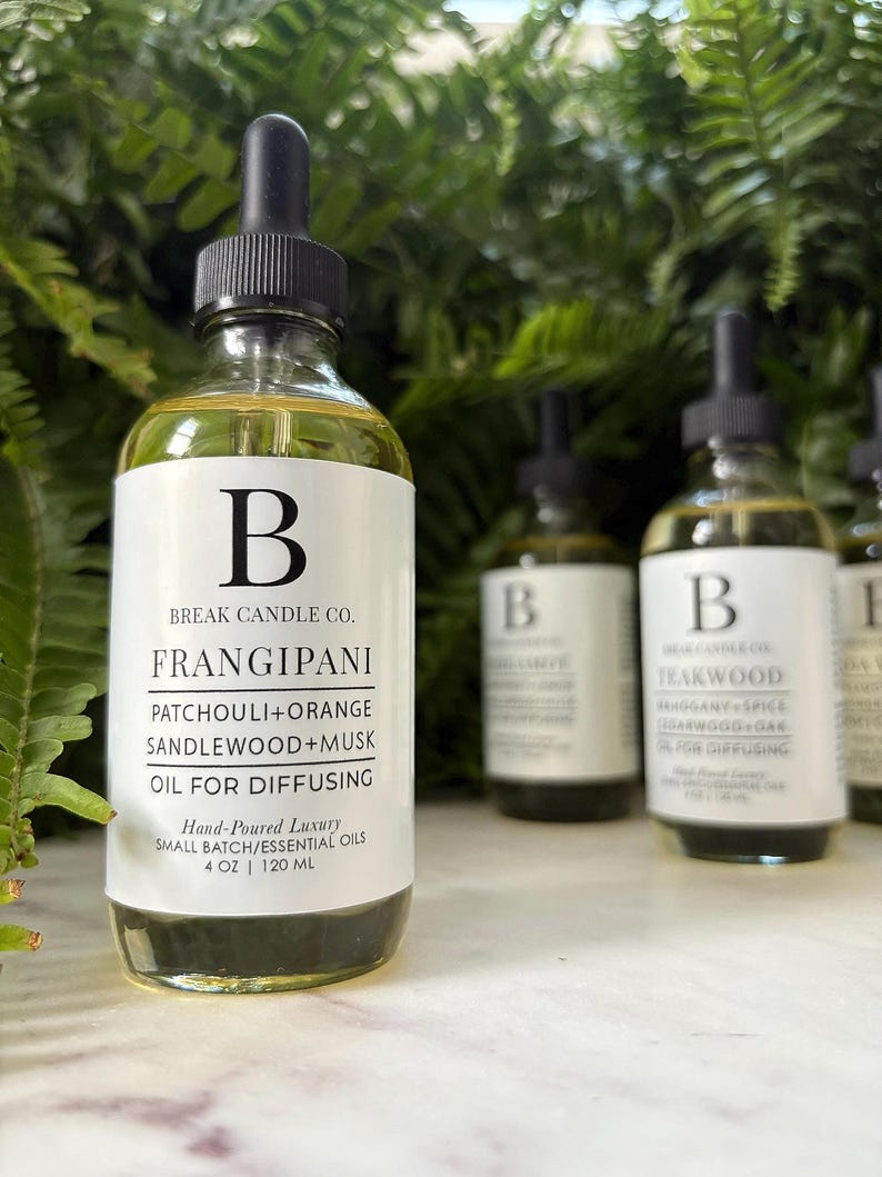 May include: Clear glass bottles of essential oil with black droppers. The labels read "FRANGIPANI" and "TEAKWOOD". The bottles are 4 oz / 120 ml. The background is a lush green plant.