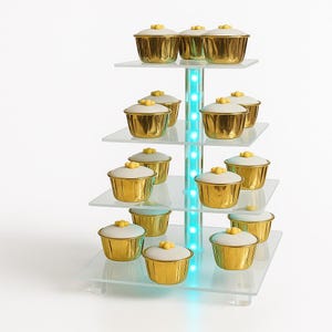 May include: A tiered dessert stand with multiple gold-colored cupcake holders, each topped with a white frosting and yellow decoration. The stand has clear glass shelves and a central column with blue LED lights.