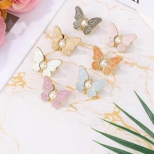 May include: A collection of butterfly-shaped brooches in various colors, including pink, blue, orange, and gold. Each brooch features a pearl in the center. The brooches are arranged on a marble-patterned surface. The text "On of the Year" is visible.