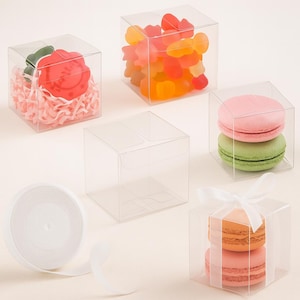 Peut inclure: Plusieurs boîtes carrées en plastique transparent contenant divers articles. Une boîte contient un biscuit rouge avec des accents verts sur du papier rose. Une autre contient des bonbons gélifiés colorés. Deux boîtes ont des macarons et une a un ruban blanc.