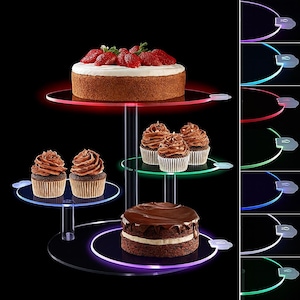 Puede incluir: Un soporte para pasteles de tres niveles de acrílico transparente con iluminación LED. El soporte exhibe un pastel con fresas, cupcakes y un pastel de chocolate. Los bordes del soporte se iluminan con luces rojas, verdes, azules y moradas.