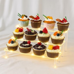 Könnte beinhalten: Eine dreistufige Präsentation von zwölf Cupcakes, jeweils in einer braunen Papierform mit goldenem Rand. Die Cupcakes sind mit weißem Zuckerguss und verschiedenen Früchten wie Erdbeeren, Blaubeeren und Mango belegt. Die Präsentation wird von kleinen, warmen Lichtern beleuchtet.