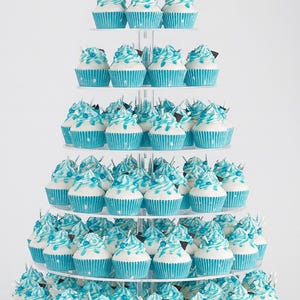 Puede incluir: Un soporte para cupcakes de varios niveles lleno de cupcakes. Cada cupcake tiene un envoltorio azul y glaseado blanco con remolinos y chispas azules. Algunos cupcakes tienen cuadrados de chocolate. El soporte es de plástico transparente.