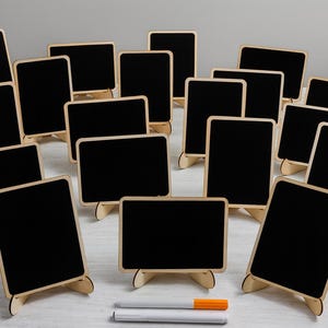 May include: A collection of small, rectangular black chalkboards with wooden frames and stands. Two white markers, one with an orange cap, are in the foreground. The chalkboards are arranged on a light-colored surface.
