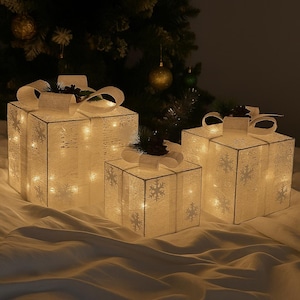 Op de afbeelding: Drie verlichte cadeauverpakkingen met witte sneeuwvlokpatronen en witte strikken. De dozen zijn van binnenuit verlicht, waardoor een warme gloed ontstaat. Op de achtergrond staat een kerstboom met ornamenten.